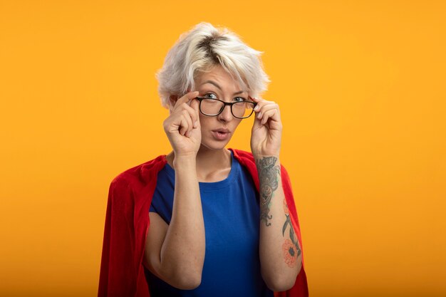 Superwoman confiada con capa roja en gafas ópticas mira al frente aislado en la pared naranja