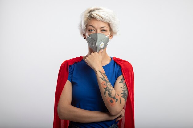 Superwoman confiada con capa roja con máscara médica pone la mano en la barbilla aislada en la pared blanca