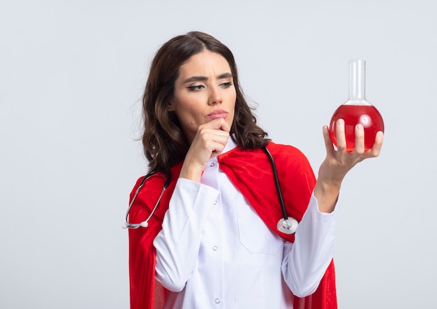 Superwoman confiada en uniforme médico con capa roja y estetoscopio pone la mano en la barbilla y mira el líquido químico rojo en un matraz de vidrio aislado en la pared blanca