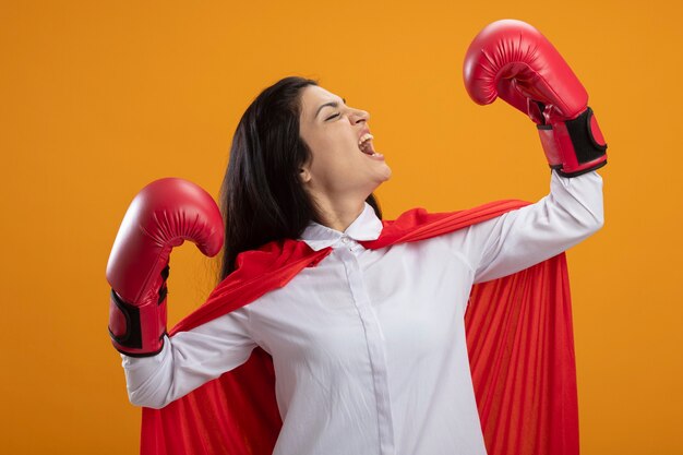 Superwoman joven confiada con guantes de boxeo haciendo un gesto fuerte girando de cabeza a lado gritando con los ojos cerrados aislados en la pared naranja