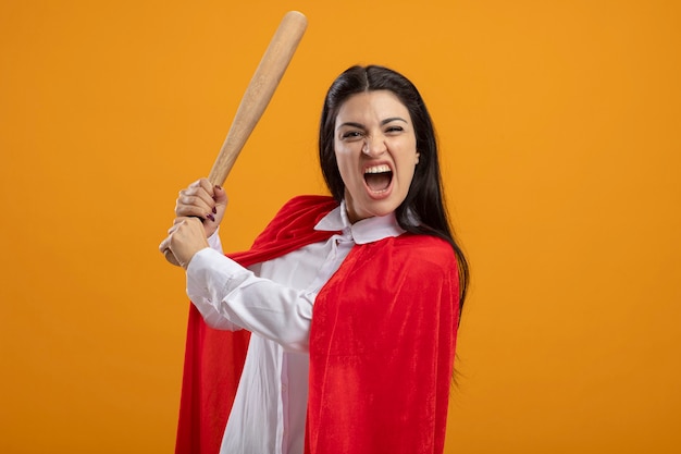 Foto gratuita superwoman joven enojada que sostiene el bate de béisbol mirando al frente aislado en la pared naranja