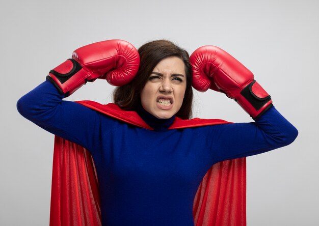 Superwoman molesta con capa roja con guantes de boxeo pone puños en la cabeza aislada en la pared blanca