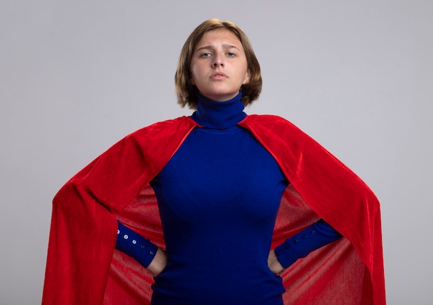 Superwoman rubia joven confiada en capa roja manteniendo las manos en la cintura mirando al frente aislado en la pared blanca