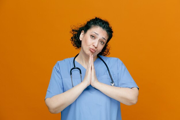 Suplicando a una doctora de mediana edad con uniforme y estetoscopio alrededor del cuello mirando a la cámara haciendo un gesto aislado en el fondo naranja