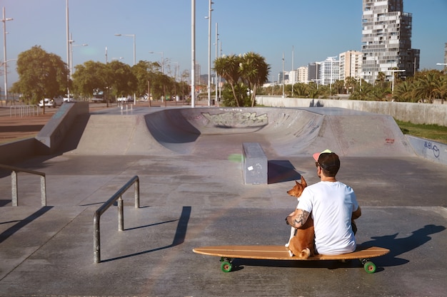 Surfer se sienta en un longboard en un skatepark de la ciudad abrazando a su perro basenji marrón y blanco, mirando el paisaje de la ciudad, disparado desde la espalda