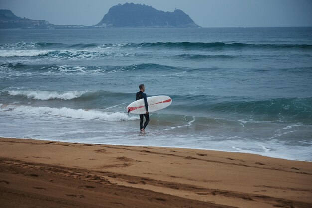 Surfista de mediana edad en traje de neopreno caminando hacia el agua en la playa de arena entre colinas