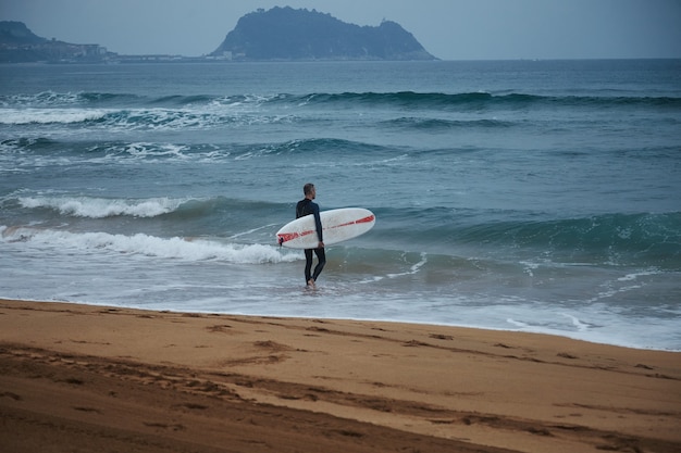 Surfista de mediana edad en traje de neopreno caminando hacia el agua en la playa de arena entre colinas