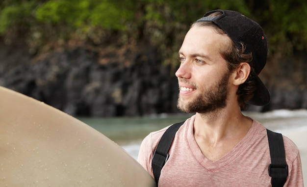 Surfista principiante joven con estilo con gorra de béisbol hacia atrás mirando al océano con una sonrisa feliz e inspirada