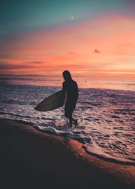 Surfista con una tabla saliendo del mar durante la puesta de sol