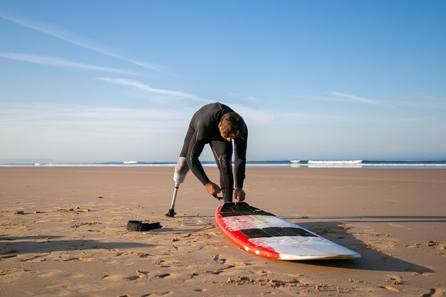 Surfista vistiendo traje de neopreno y una extremidad artificial, atando la tabla a su tobillo sobre arena.