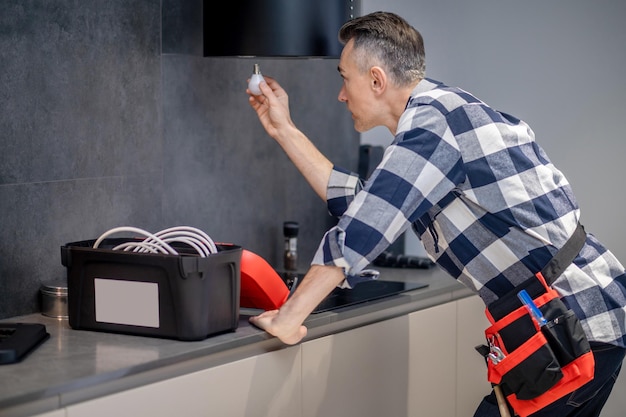 Foto gratuita sustitución de la lámpara vista posterior y lateral del hombre con camisa a cuadros con lámpara en la mano mirando la cocina