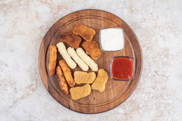 Tabla de aperitivos de madera con nuggets de pollo, palitos de queso, salchichas a la parrilla y salsas alrededor.