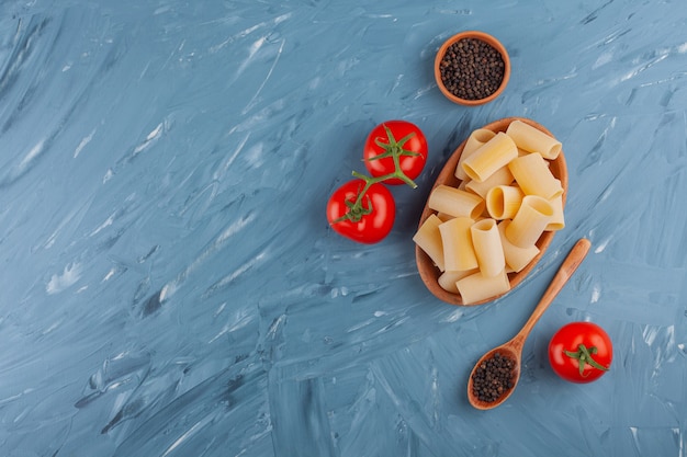 Una tabla de arcilla de pasta seca de tubo crudo con tomates rojos frescos y granos de pimienta.