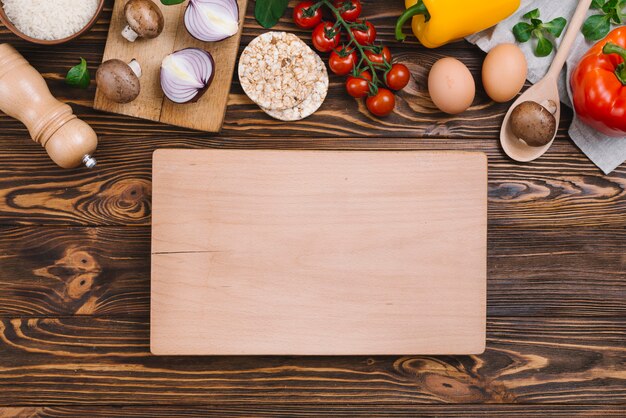 Tabla de cortar de madera en blanco con verduras crudas y pastel de arroz inflado sobre mesa de madera