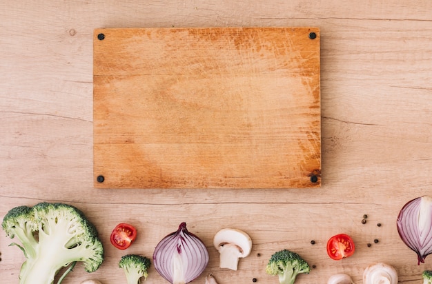 Tabla de cortar de madera con brócoli; los tomates cebolla; Champiñones y pimienta negra sobre mesa.