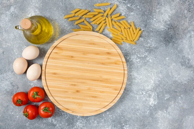 Tabla de madera de espaguetis crudos secos, huevos y tomates en la superficie de mármol.