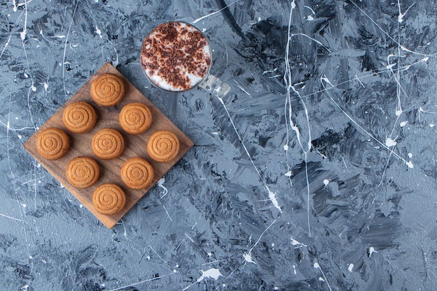 Una tabla de madera de galletas redondas dulces con una taza de cristal de sabroso café caliente sobre un fondo de mármol.