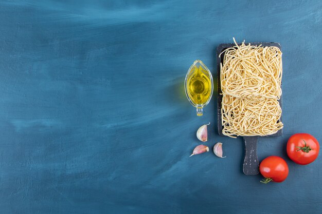 Una tabla de madera negra de fideos crudos con dos tomates rojos frescos y aceite sobre un fondo azul.