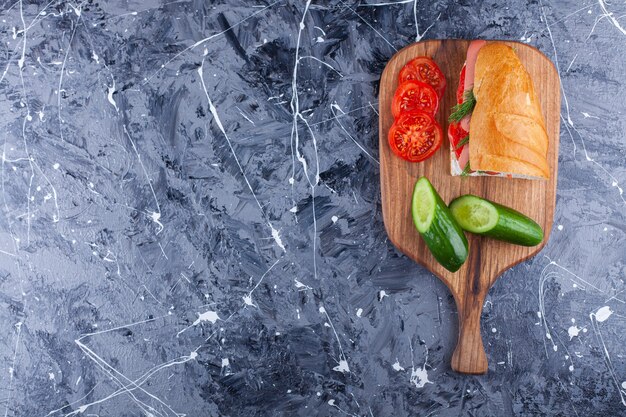 Tabla de madera de sándwich casero y verduras en rodajas sobre superficie de mármol.