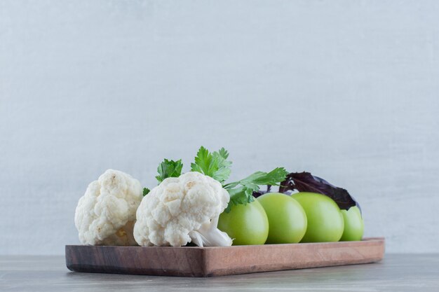 Tabla de madera de tomates verdes y trozos de coliflor rematada con hojas de amaranto y perejil sobre mármol.