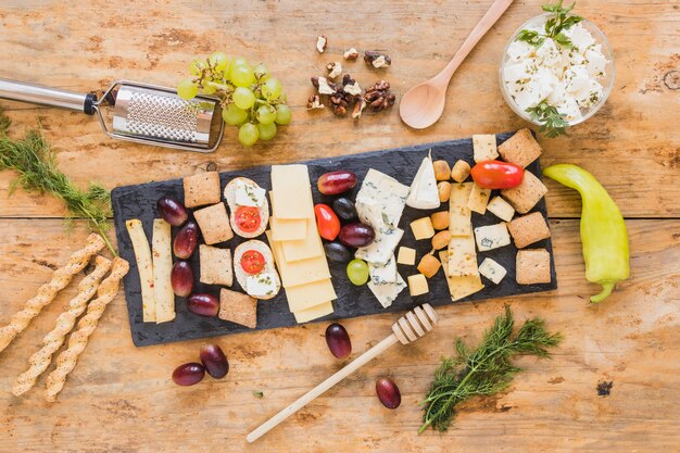 Tabla de quesos con perejil, uvas; cucharón de miel; Palitos de pan y ají verde en superficie de madera