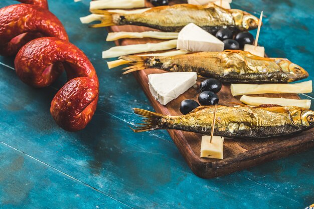 Tablero de aperitivos con rodajas de salchicha, cubitos de queso y aceitunas negras con galletas y pescado seco en la mesa azul