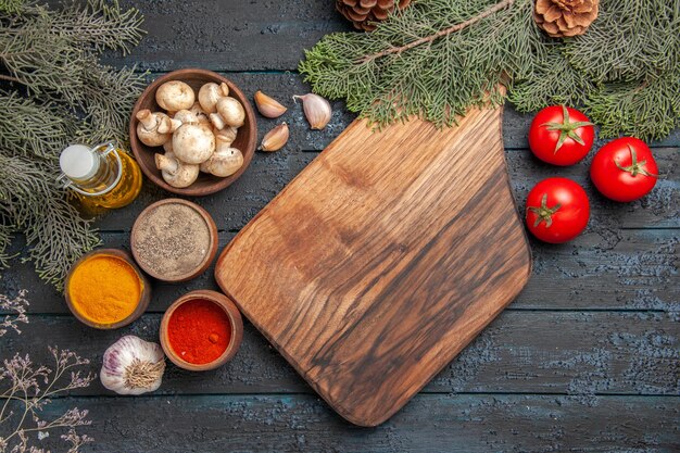 Tablero de vista superior y especias Tabla de cortar marrón de madera junto a un tenedor ajo especias coloridas aceite en botella tres tomates y tazón de champiñones debajo de ramas con conos