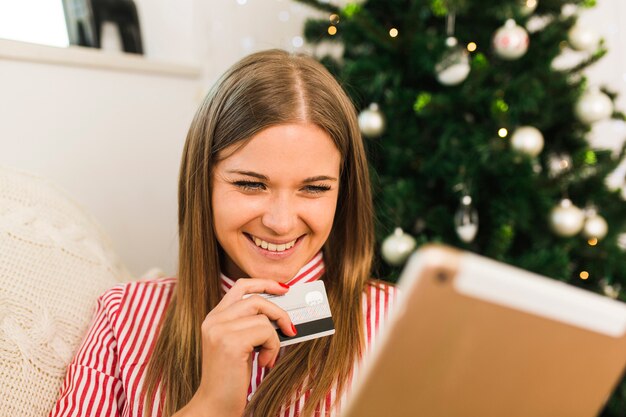 Tableta alegre de la tenencia de la señora y tarjeta de crédito cerca del árbol de navidad
