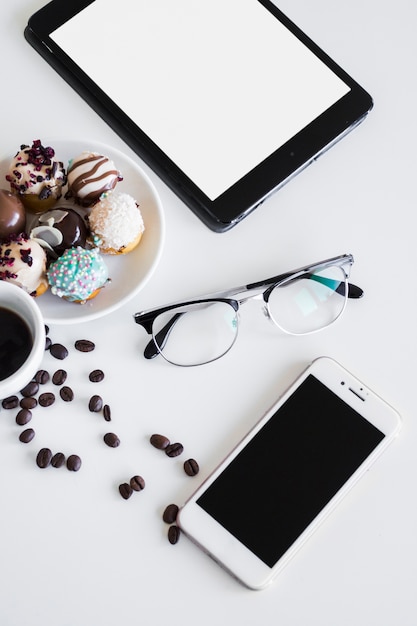 Tableta cerca de teléfono inteligente, taza, gafas y galletas en el plato