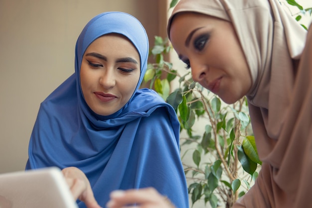 Tableta de desplazamiento. Hermosas mujeres árabes reunidas en café o restaurante, amigos o reunión de negocios. Pasar tiempo juntos, hablando, riendo. estilo de vida musulmán. Modelos elegantes y felices con maquillaje.