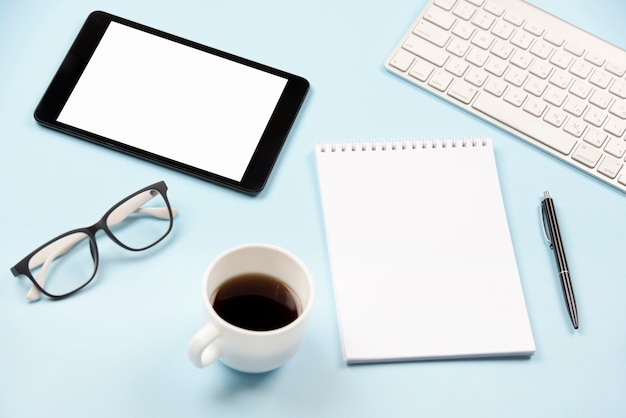 Tableta digital; los anteojos; taza de café; libreta espiral en blanco; pluma y teclado sobre fondo azul