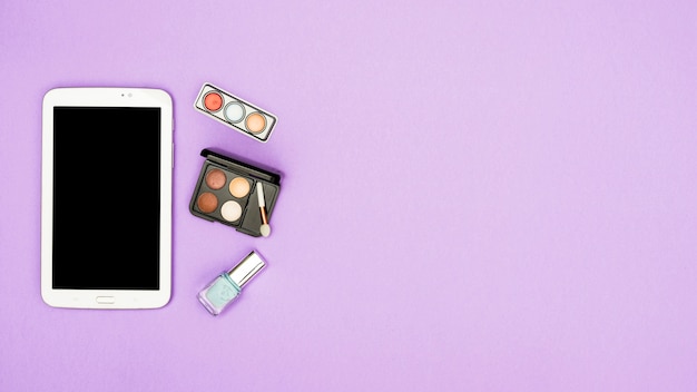 Tableta digital con paleta de sombra de ojos y botella de esmalte de uñas sobre fondo morado