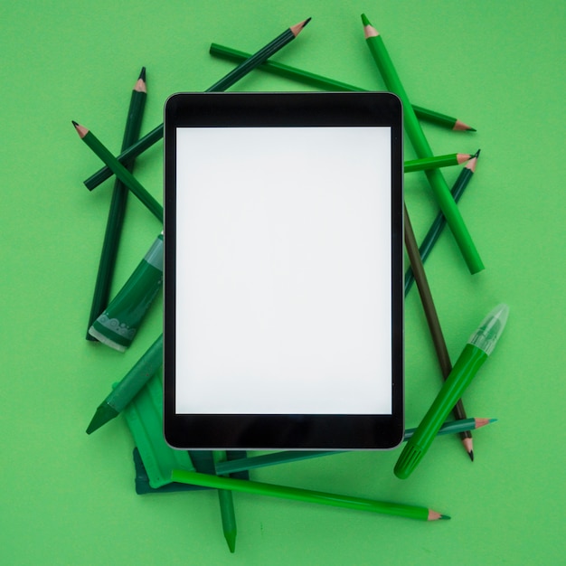 Foto gratuita tableta digital de primer plano con pantalla en blanco sobre lápices de colores; arcilla; tubo y marcador sobre fondo verde oscuro