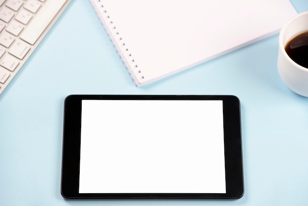 Tableta digital con teclado; Bloc de notas espiral y taza de café sobre fondo azul