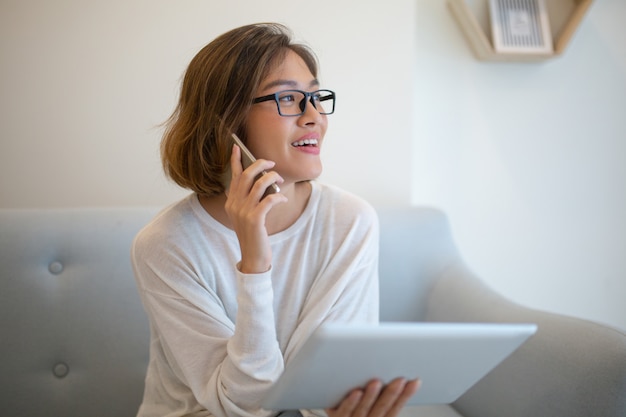 Tableta sonriente de la tenencia de la mujer y hablar en el teléfono en el sofá