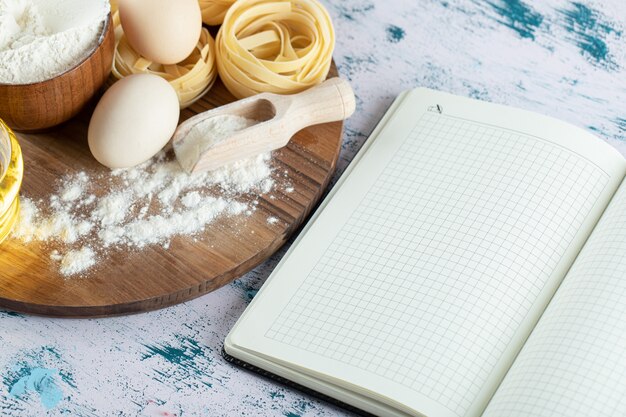 Tagliatelle con aceite, huevo y tazón de harina sobre tabla de madera y cuaderno.