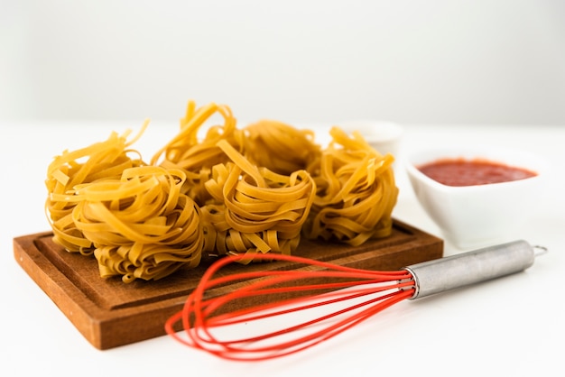 Tagliatelle pasta sobre tabla para cortar madera con batidor rojo sobre fondo blanco.