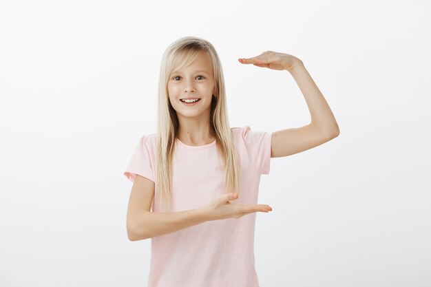 El tamaño es enorme. Filmación en interiores de fascinada niña europea adorable satisfecha con cabello rubio, dando forma a una cosa larga con palmas y sonriendo ampliamente, explicando y mostrando la forma del espacio de la copia sobre una pared gris