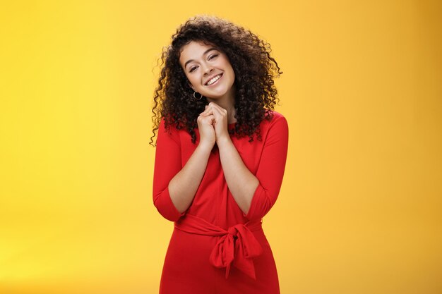 Tan dulce te amo. Retrato de tierna y tonta linda mujer joven de pelo rizado en vestido rojo inclinando la cabeza cogidos de la mano cerca de la cara y sonriendo tocado durante el conmovedor momento de la confesión.