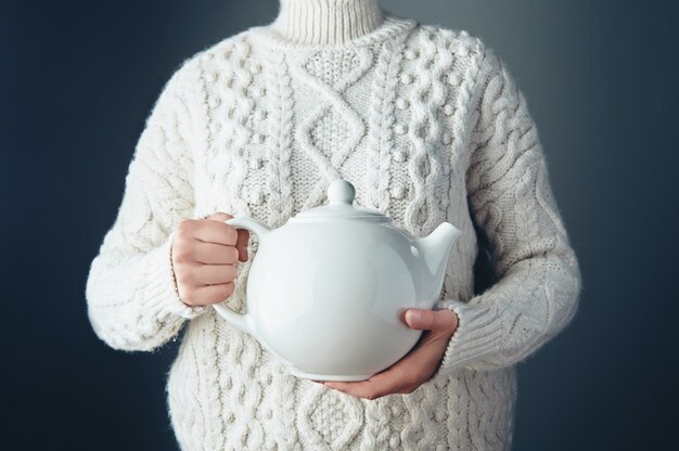 Tapot blanco grande con té en las manos en el aire. Mujer irreconocible vestía suéter blanco de punto grueso. Vista frontal.