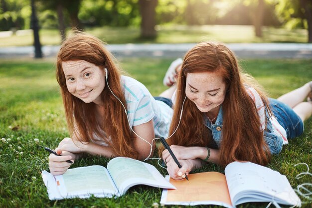 Tarea, ¿me estás tomando el pelo? Dos chicas encantadoras con el pelo rojo tumbadas y relajadas en el césped durante el tiempo libre haciendo la tarea. Hermana ayudando a su hermano con lecciones sonriendo y riendo. Copiar espacio.