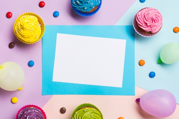 Tarjeta azul y blanca en blanco rodeada de globo; muffins y gemas sobre fondo coloreado