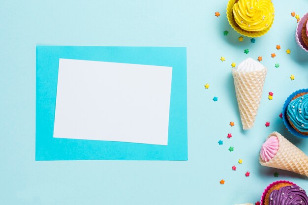 Tarjeta de felicitación en blanco cerca de los sprinkles; Conos de gofres y muffins sobre fondo azul