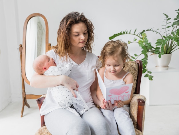 Tarjeta de felicitación de la lectura de la madre y de la hija junto que se sienta en la silla del brazo en el bebé casero de la explotación agrícola