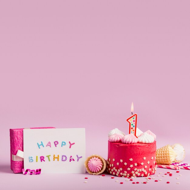 Tarjeta de feliz cumpleaños cerca del pastel con velas encendidas y caja de regalo sobre fondo púrpura