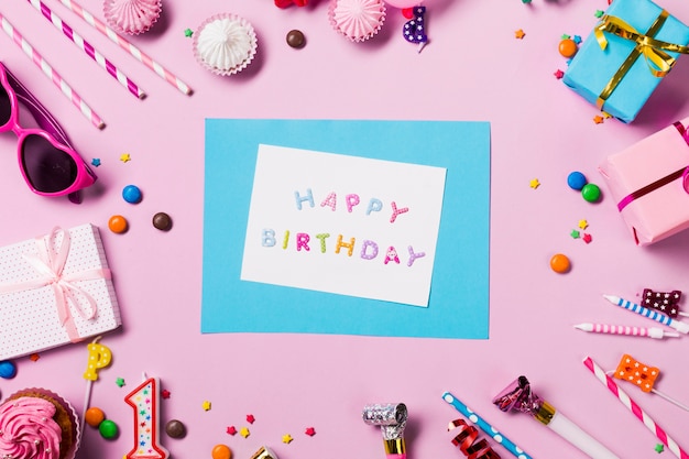 Tarjeta de feliz cumpleaños rodeada de artículos de cumpleaños sobre fondo rosa