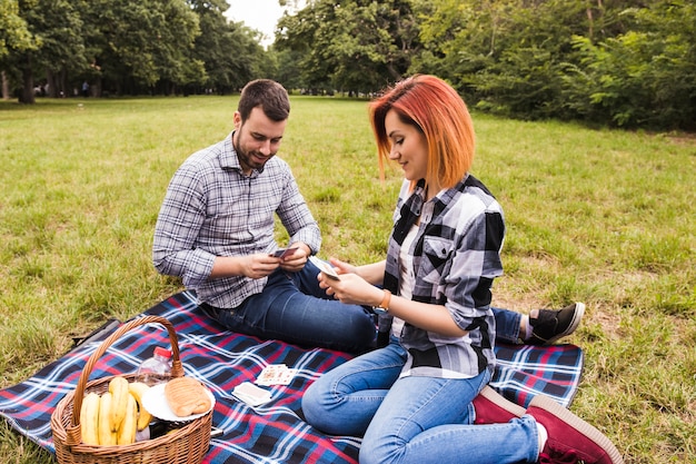 Tarjetas que juegan jovenes sonrientes de los pares que se sientan en la manta en la comida campestre