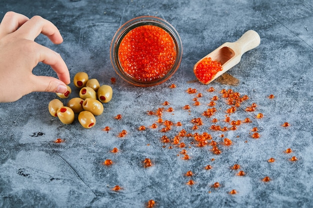 Un tarro de caviar rojo y aceitunas verdes sobre fondo de mármol.