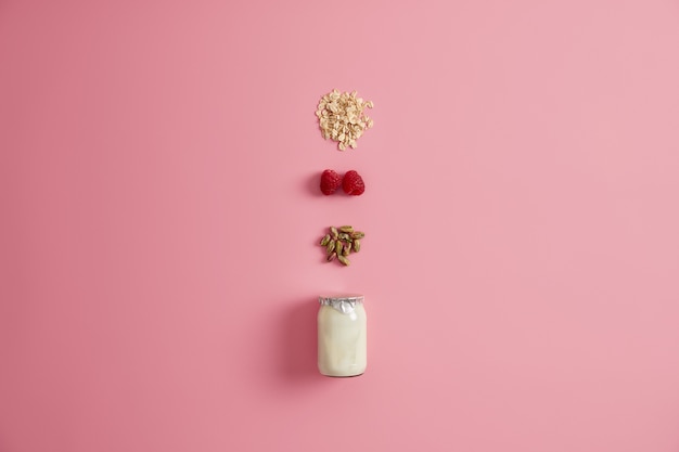 Tarro de cristal con yogur, pistacho, frambuesa roja y cereales para mezclar y comer. Fondo rosa. Desayuno de dieta saludable. Ingredientes naturales para papilla o snack rápido. Comida vegetariana