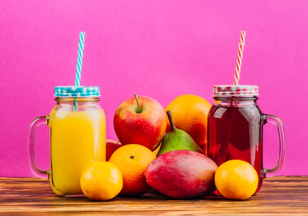 Tarros de albañil de jugo rojo y amarillo con pajitas y frutas frescas sobre fondo rosa